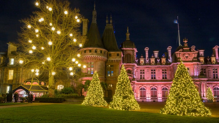 A view of Waddesdon Manor illuminated as part of a Christmas lights event, with three lit-up Christmas trees in front of the manor and a large tree hung with bauble lights to the left
